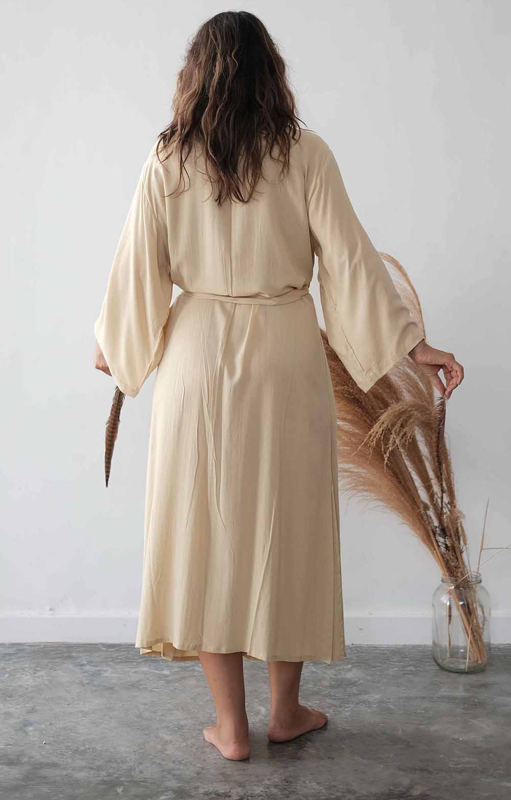Woman wearing a beige robe holding dried plants against a white wall.