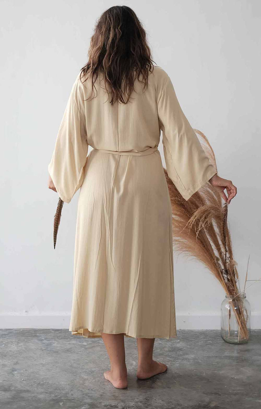 Woman wearing a beige robe holding dried plants against a white wall.