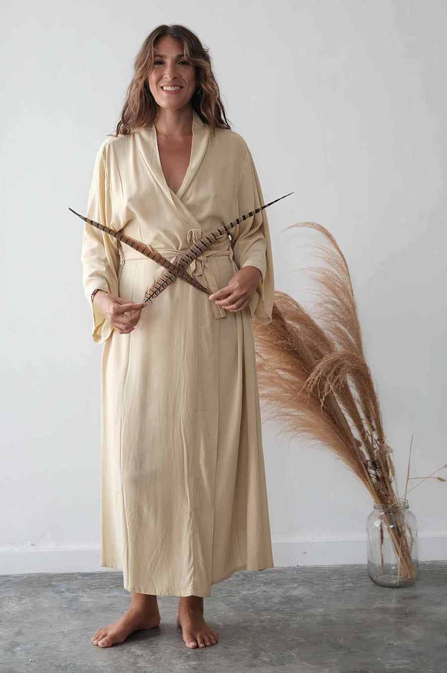 Woman in a beige robe holding dried plants against a white wall.