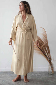 Woman wearing a beige robe holding dried pampas grass against a white wall.