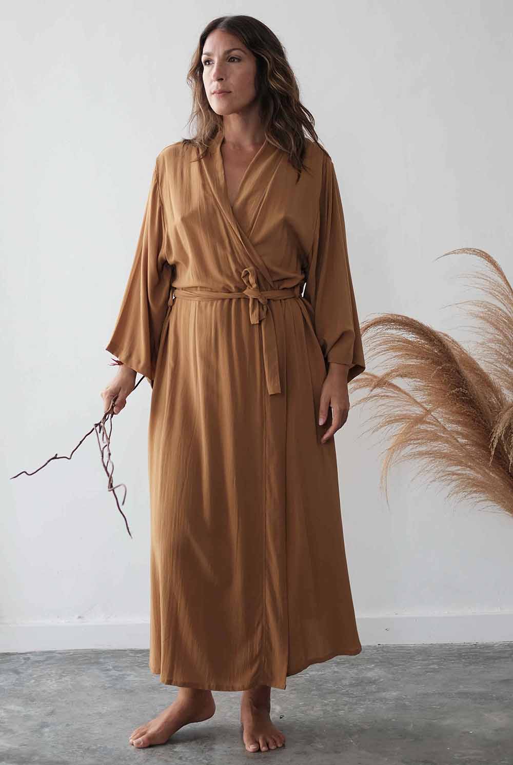 Woman wearing a long brown robe holding a branch against a white wall.