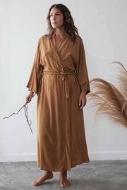 Woman wearing a long brown robe holding a branch against a white wall.