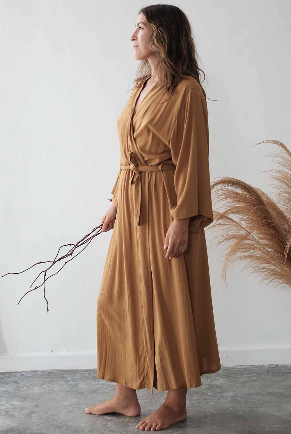 Woman wearing a long mustard yellow dress holding a branch against a white wall.