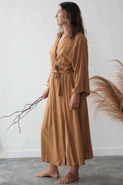 Woman wearing a long mustard yellow dress holding a branch against a white wall.