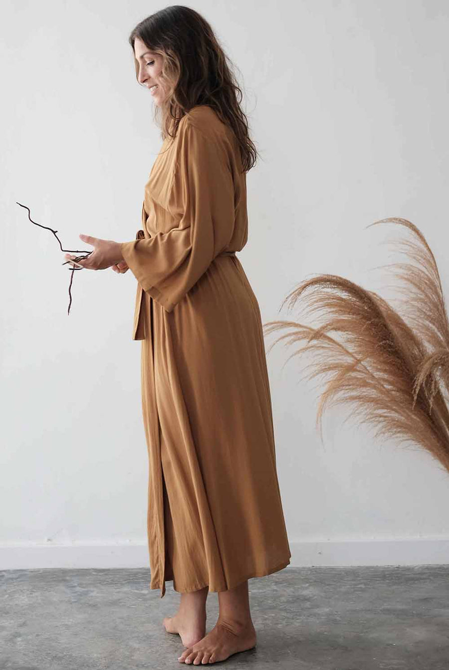 Woman wearing a long brown dress holding a branch against a white wall with pampas grass.