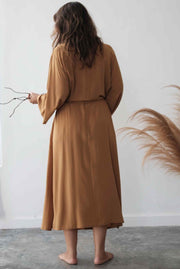 Woman wearing a long brown dress standing against a white wall with decorative elements.
