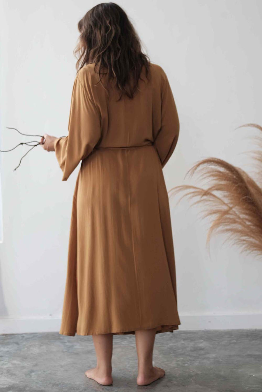 Woman wearing a long brown dress standing against a white wall with decorative elements.