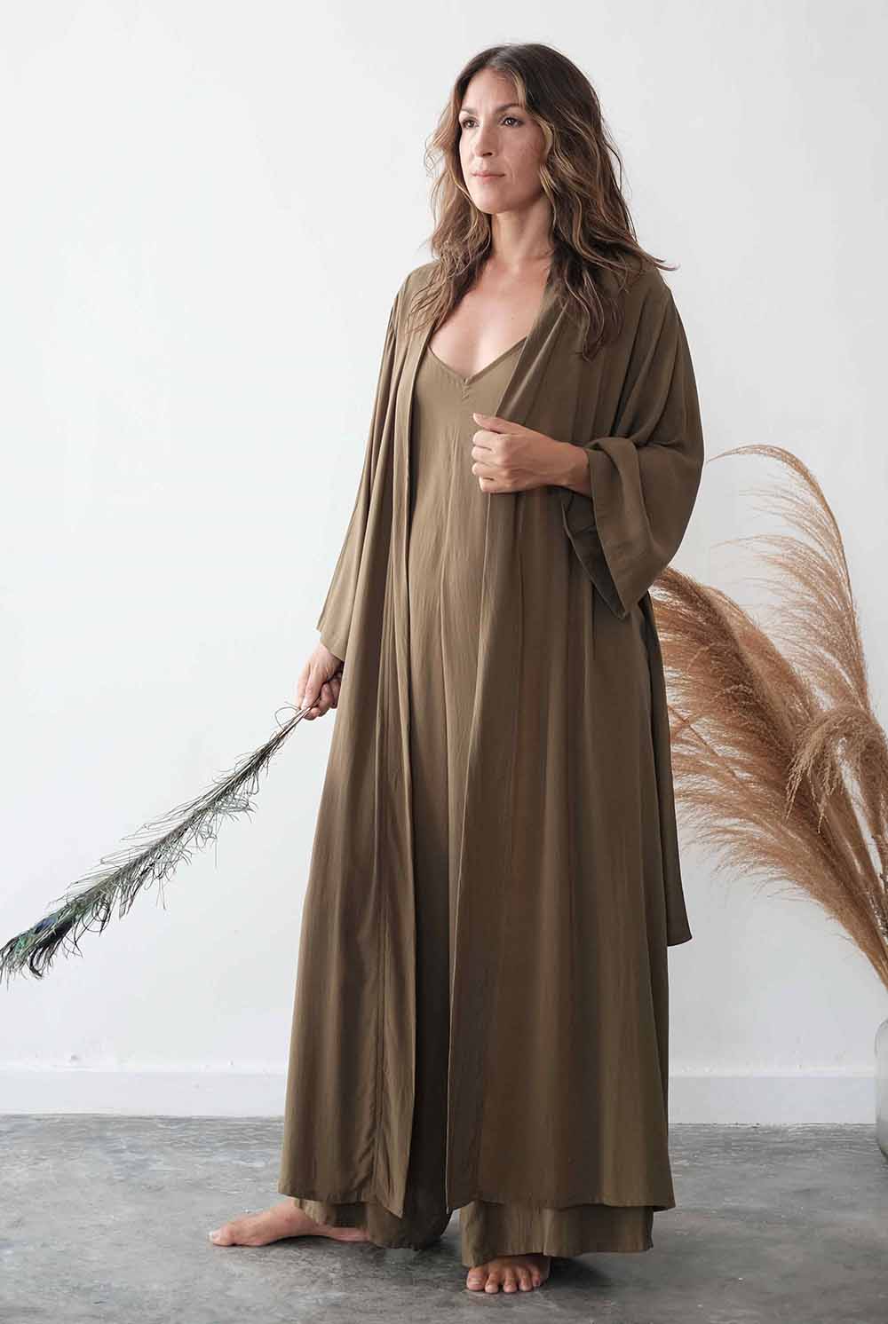 Woman wearing a long brown dress holding a dried plant against a white wall.