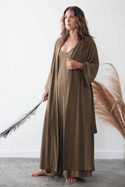 Woman wearing a long brown dress holding a dried plant against a white wall.