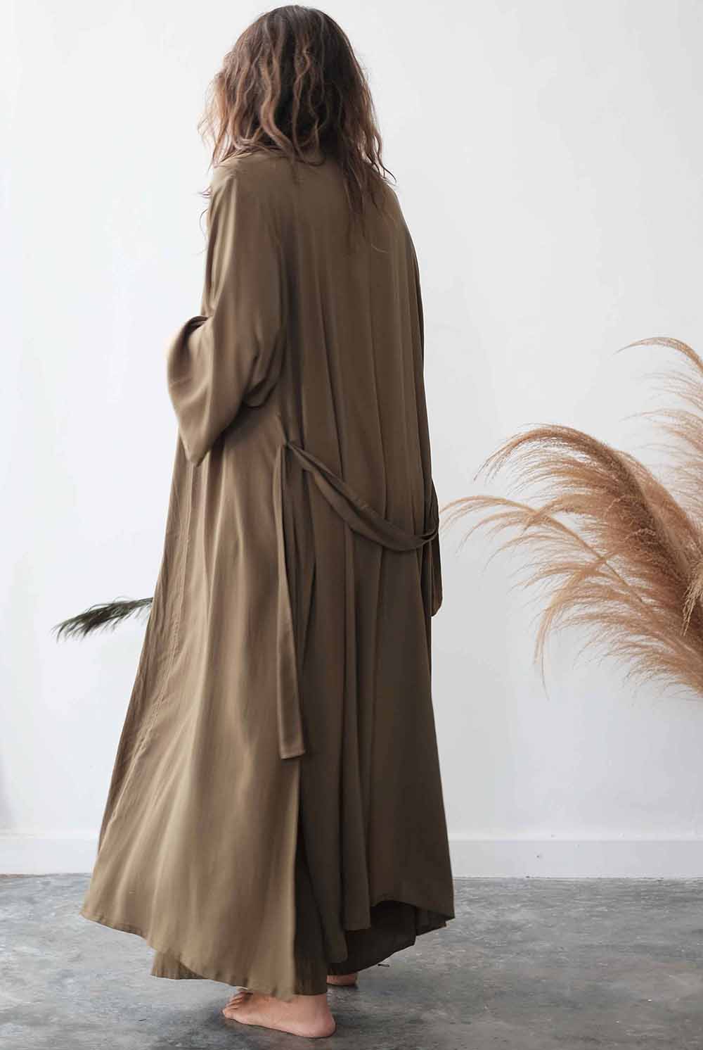 Person wearing a long, light brown robe standing against a white wall with dried pampas grass.