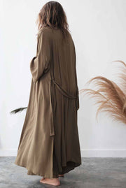 Person wearing a long, light brown robe standing against a white wall with dried pampas grass.
