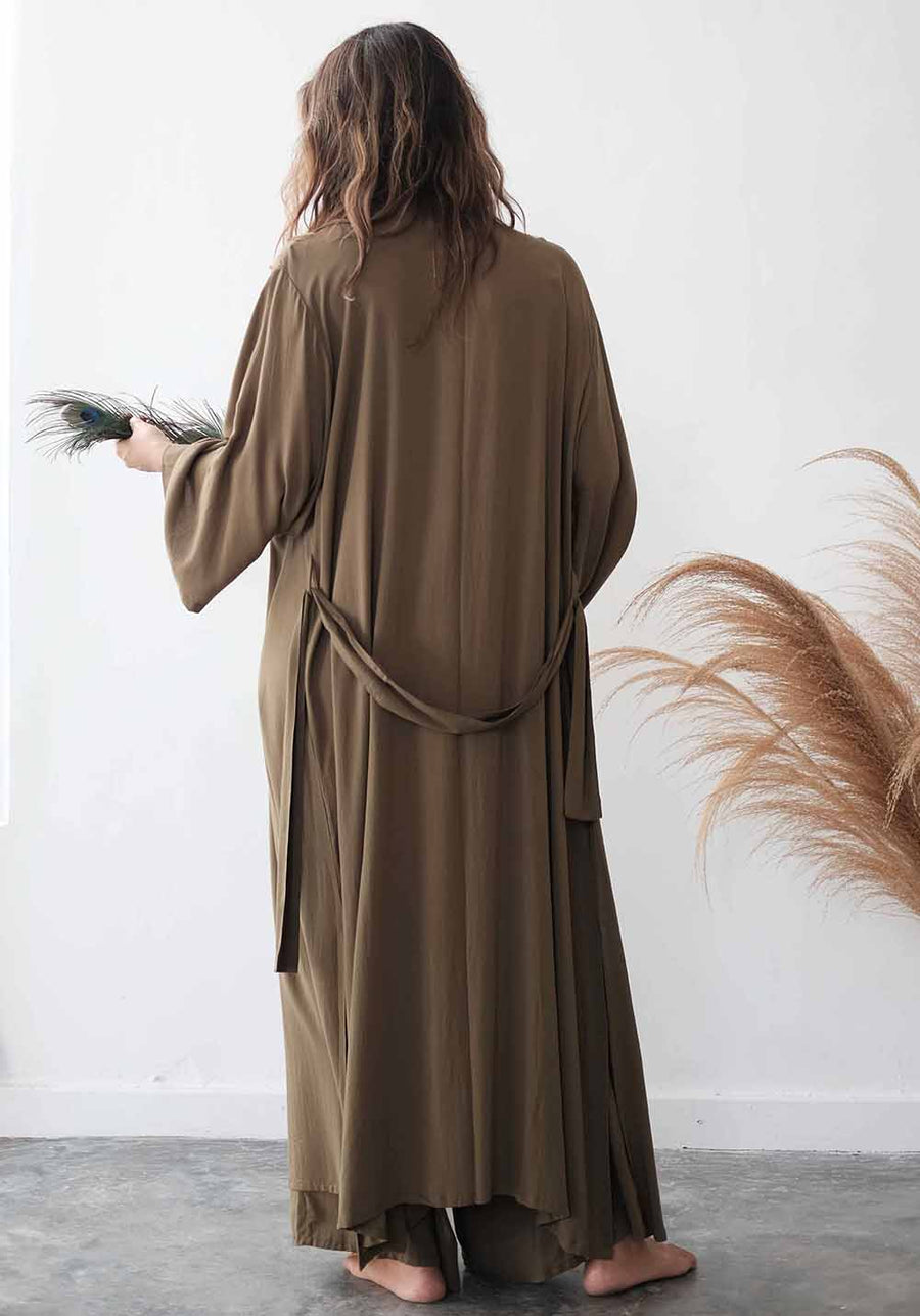 Person wearing a long brown dress holding feathers against a white wall with decorative elements.