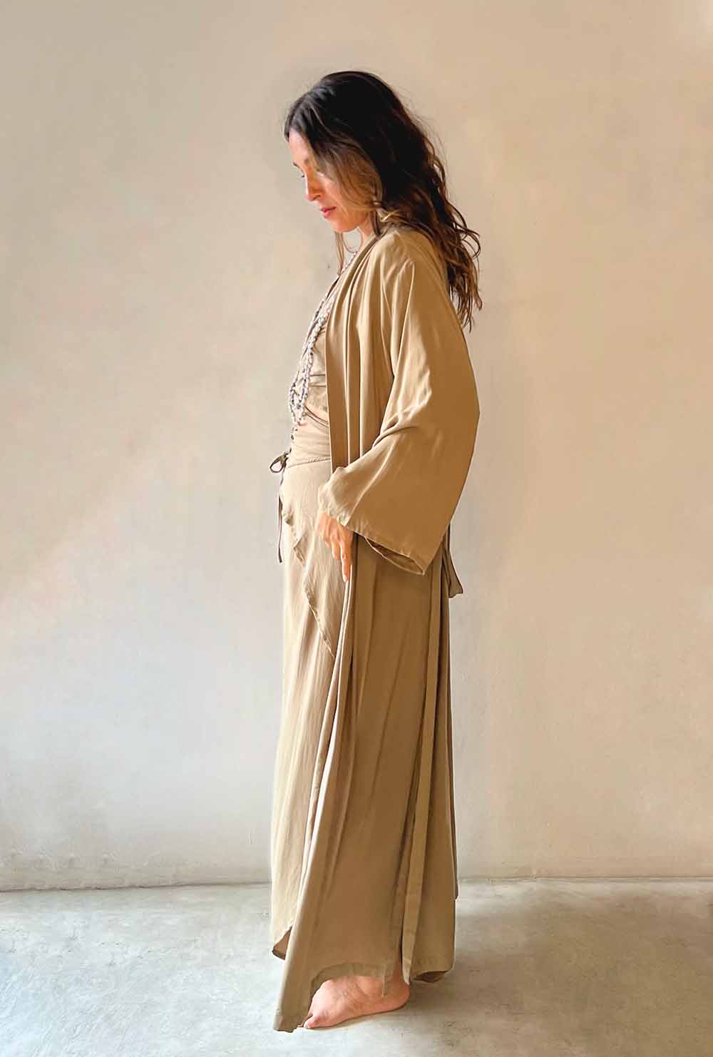 Woman wearing a beige robe standing against a plain wall.