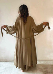 Person wearing a long beige dress holding a belt against a plain background