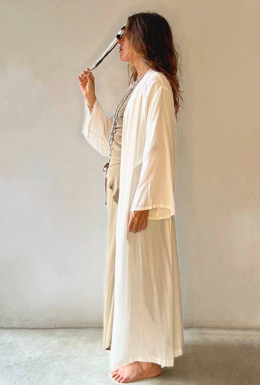 Woman in a long beige robe holding a pen against a plain wall.
