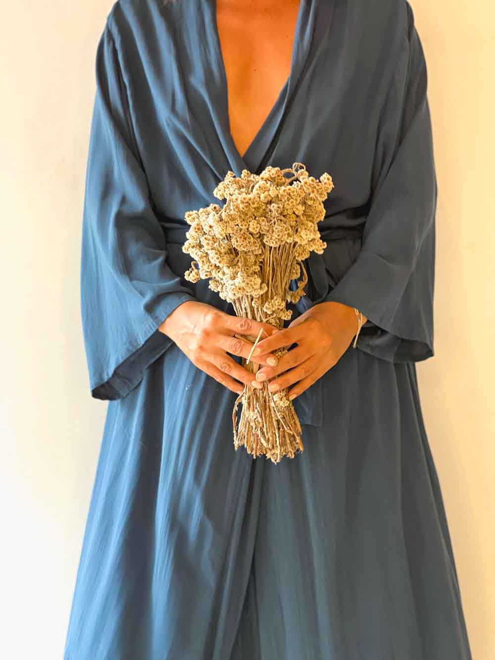 Person wearing a long, dark blue dress holding a bouquet of dried flowers against a beige background