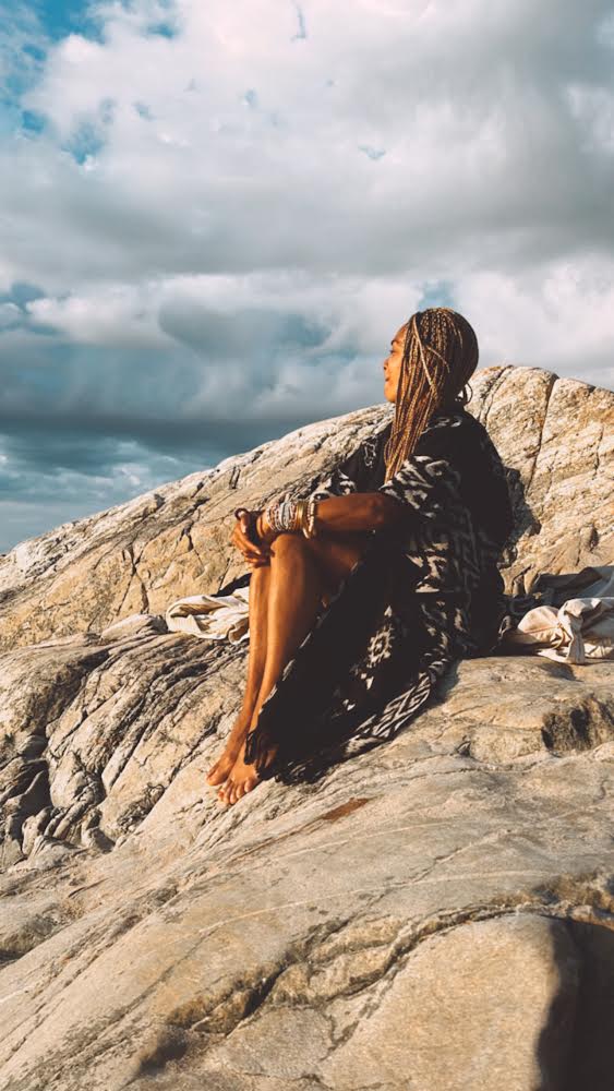 A person sitting on rocky terrain wearing a black kaftan with a traditional Ikat pattern, paired with a matching belt.