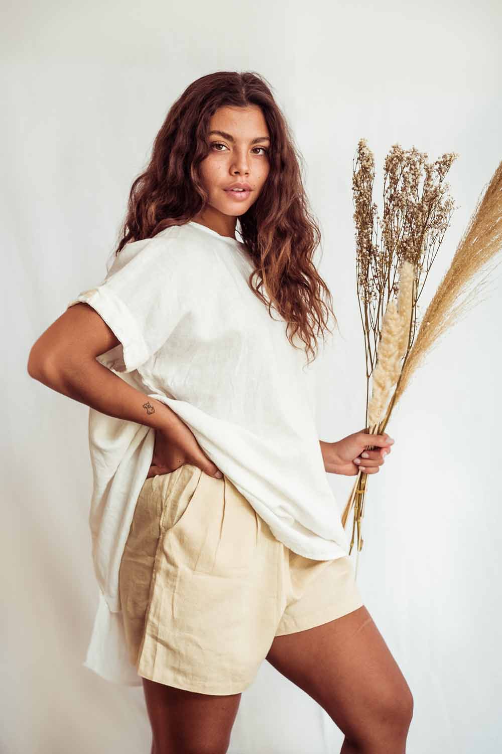 Woman wearing a white top and beige shorts holding dried plants against a plain background