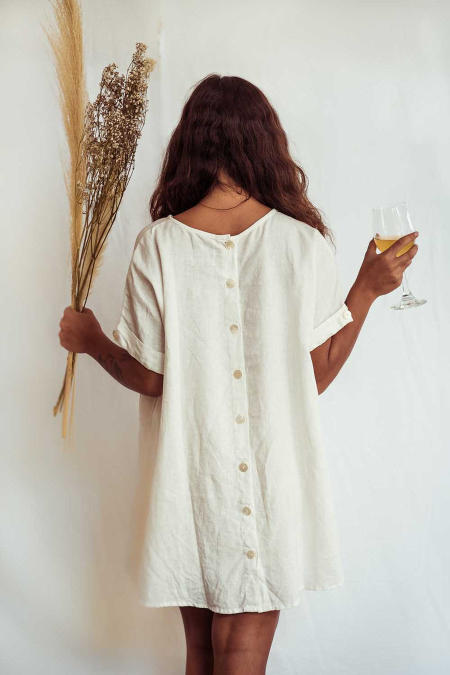 Woman in a white dress holding dried plants and a glass of wine against a white background