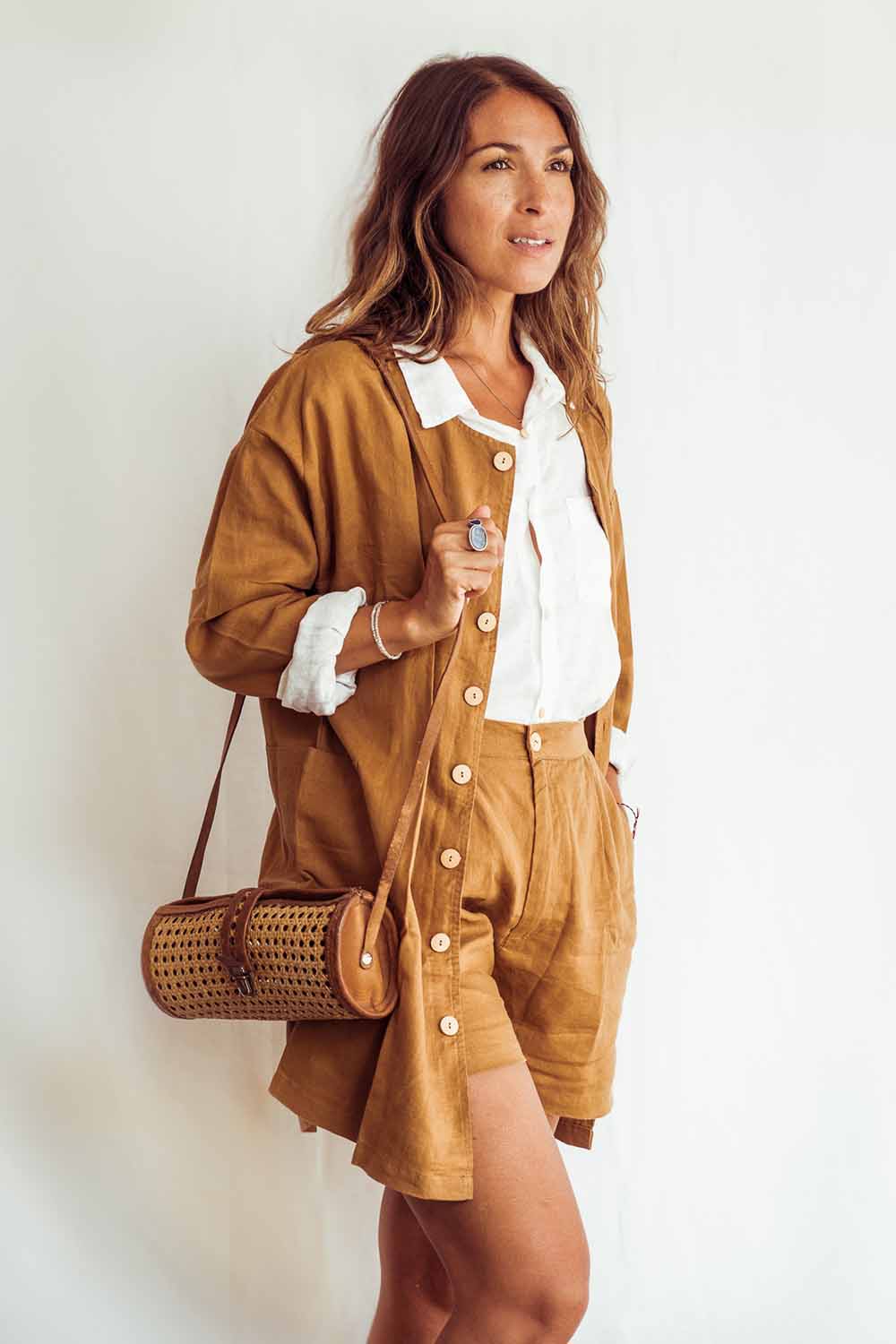 Woman wearing a brown outfit with a white shirt, holding a woven bag against a white background