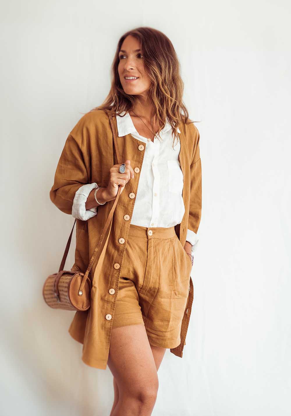 Woman wearing a brown jacket and shorts with a white shirt, holding a woven bag against a white background.
