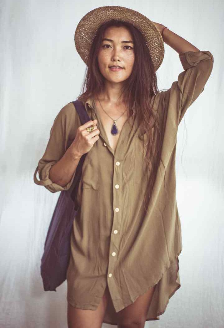 A woman wearing a beige shirt dress with buttons down the front, styled as an open shirt. She has accessorized with a wide-brimmed hat, a necklace, and a handbag.