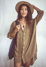 A woman wearing a beige shirt dress with buttons down the front, styled as an open shirt. She has accessorized with a wide-brimmed hat, a necklace, and a handbag.