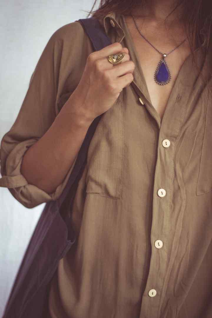 A woman wearing a beige shirt dress with buttons down the front, styled as an open shirt. She has accessorized with a wide-brimmed hat, a necklace, and a handbag.