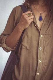 A woman wearing a beige shirt dress with buttons down the front, styled as an open shirt. She has accessorized with a wide-brimmed hat, a necklace, and a handbag.