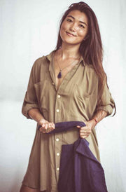 A woman wearing a beige shirt dress with buttons down the front, styled as an open shirt. She has accessorized with a wide-brimmed hat, a necklace, and a handbag.