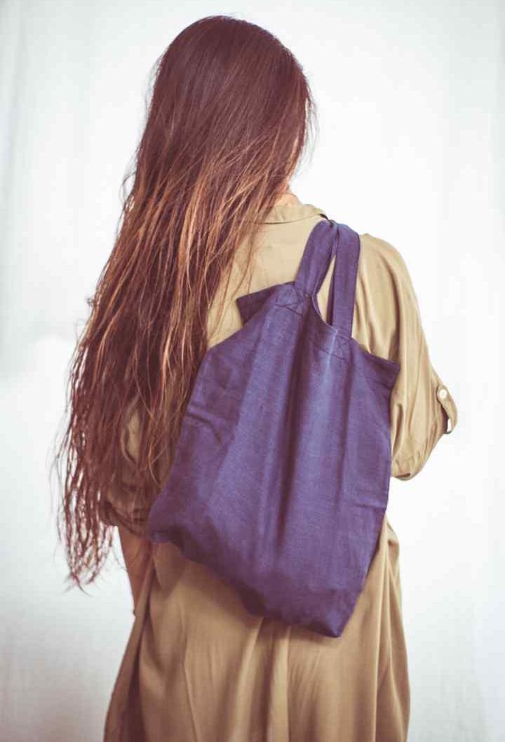 A woman wearing a beige shirt dress with buttons down the front, styled as an open shirt. She has accessorized with a wide-brimmed hat, a necklace, and a handbag.