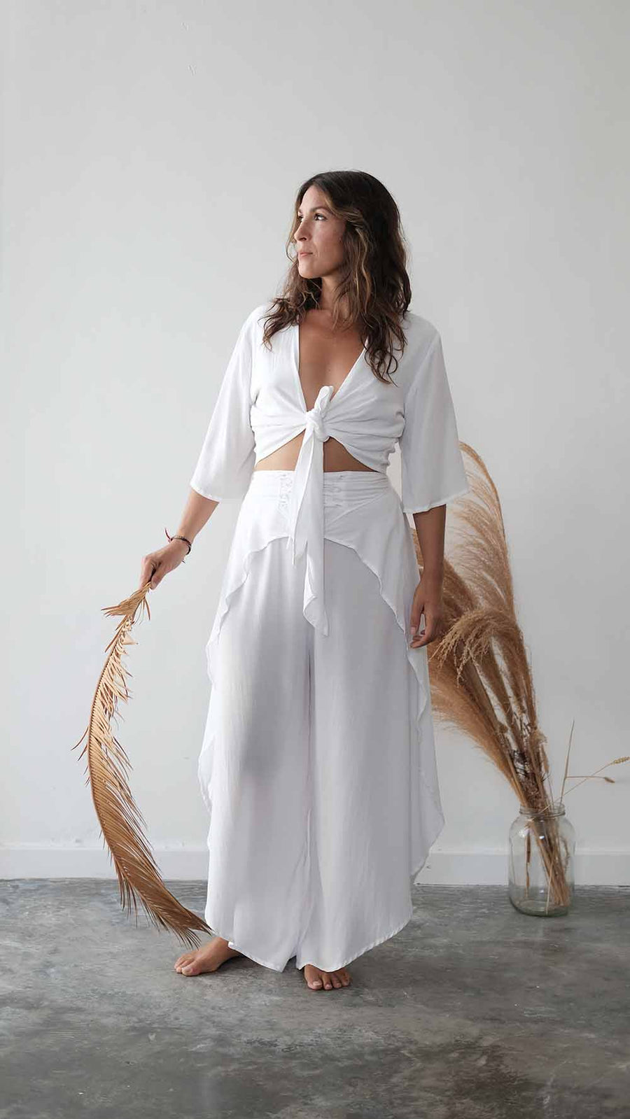 Woman wearing a white outfit standing with dried plants against a plain background