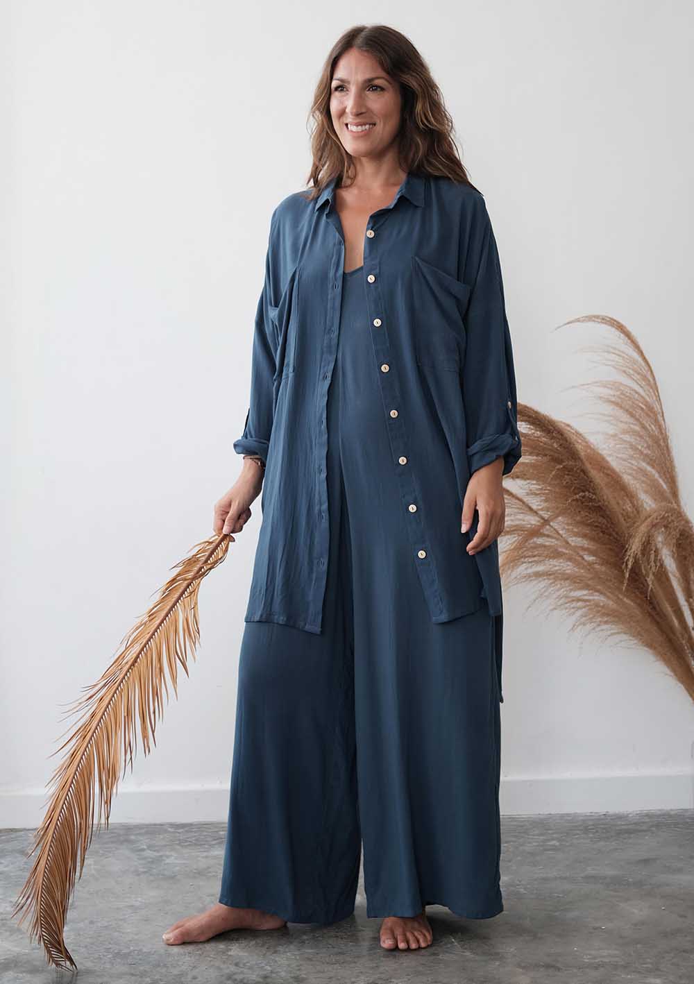 Woman wearing a blue pajama set holding dried pampas grass against a white wall.