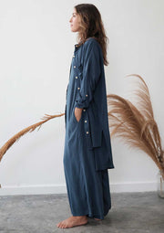 Woman wearing a long blue dress with white buttons against a white wall.