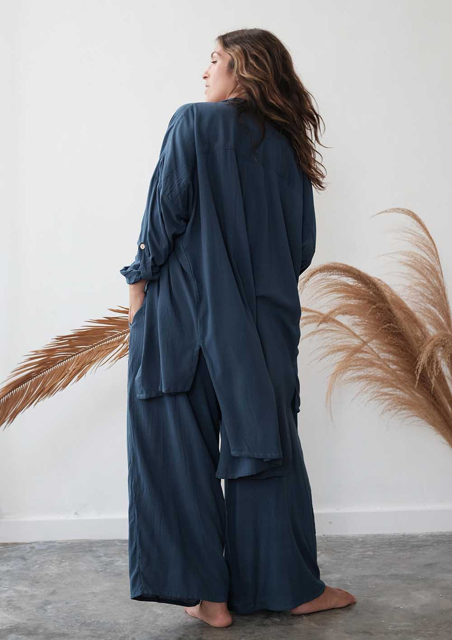 Woman wearing a dark blue outfit standing against a white wall with dried plants.