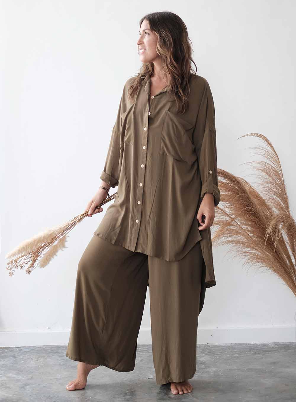Woman wearing a brown outfit holding dried pampas grass against a white wall.