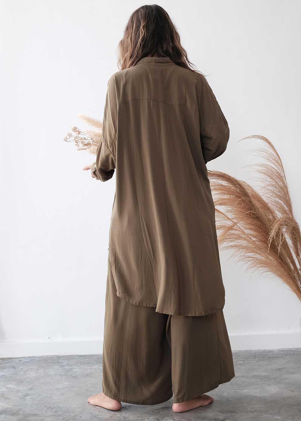 Person wearing a long brown garment standing against a white wall with dried plants.