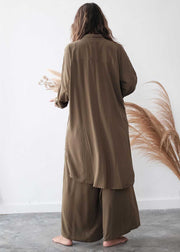 Person wearing a long brown garment standing against a white wall with dried plants.
