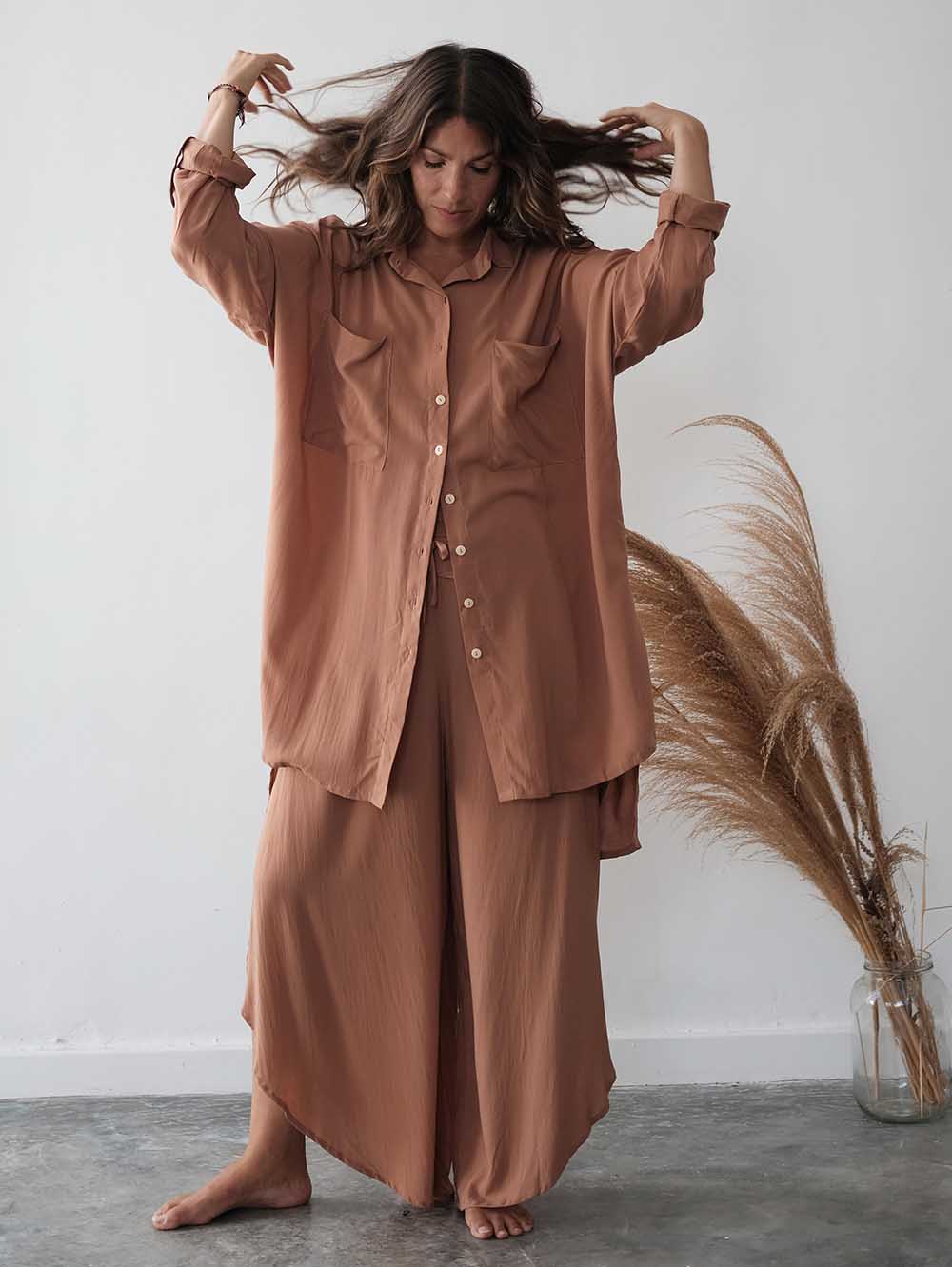Woman in a brown outfit standing next to pampas grass against a white wall