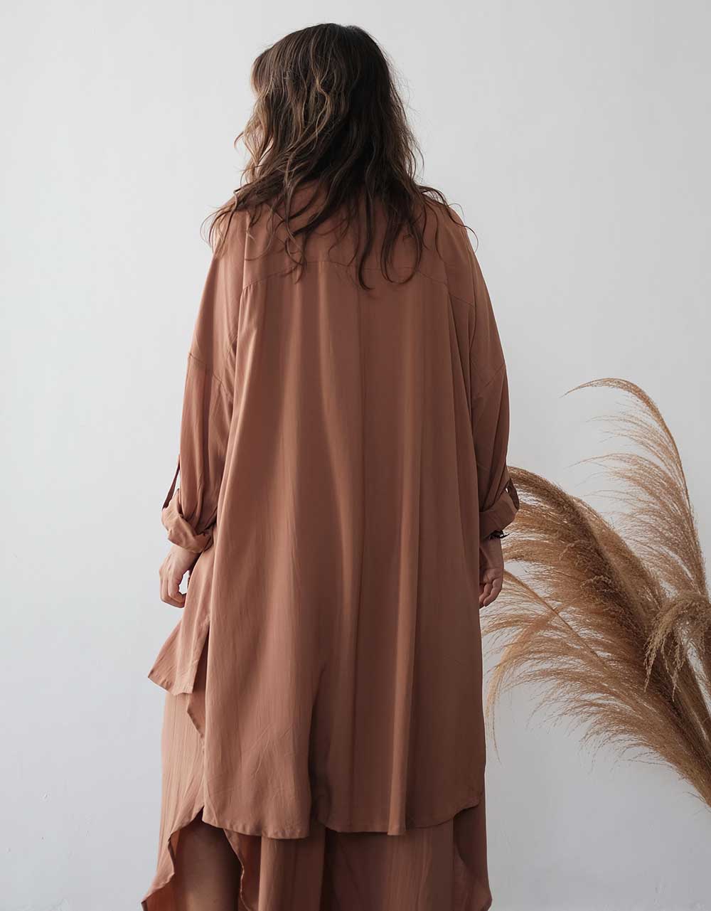 Person wearing a long brown dress standing against a white wall with dried pampas grass.
