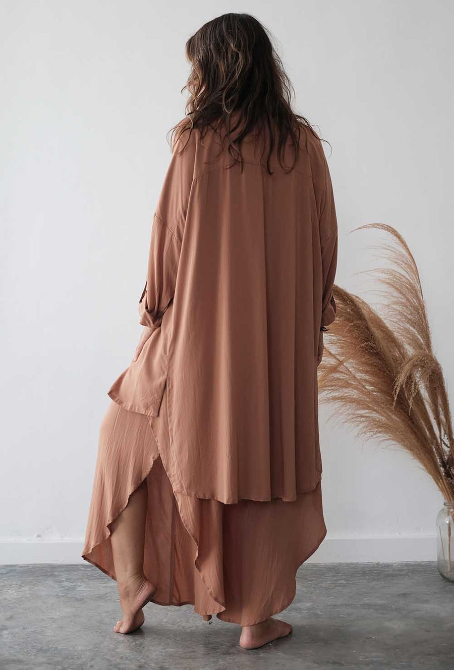 Woman wearing a long, light brown dress standing against a white wall with dried pampas grass.