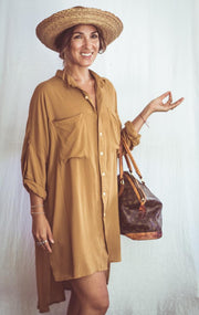 A person wearing a rust-colored, long shirt dress with buttons down the front and two chest pockets. They are also wearing a wide-brimmed hat and carrying a bag.