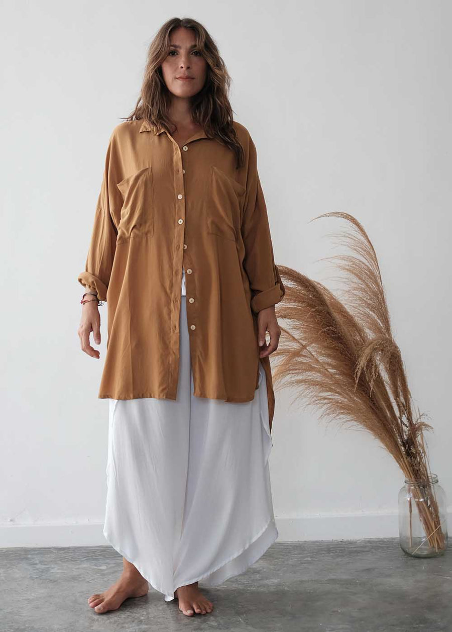 Woman wearing a brown shirt and white pants standing next to dried pampas grass against a white wall.