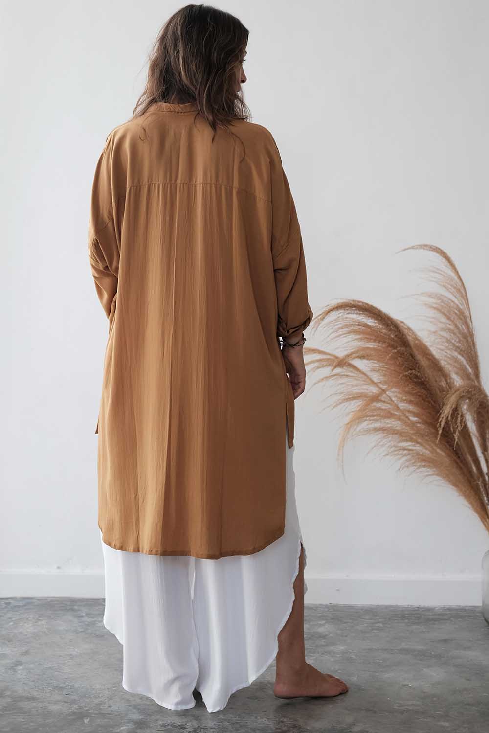 Person wearing a long brown garment over a white skirt against a white wall with dried plants.