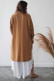 Person wearing a long brown garment over a white skirt against a white wall with dried plants.