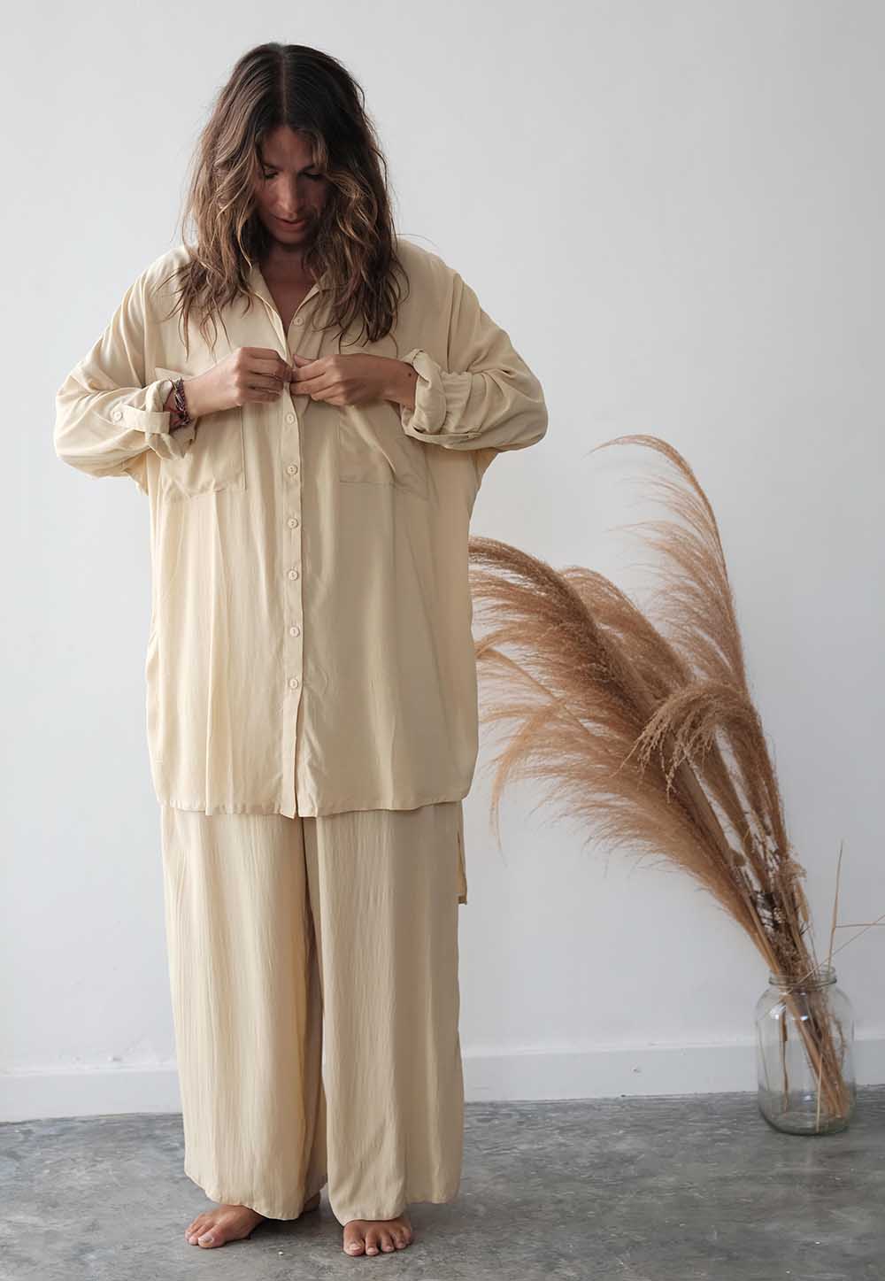 Woman wearing a beige pajama set standing next to a vase with dried pampas grass against a white wall.