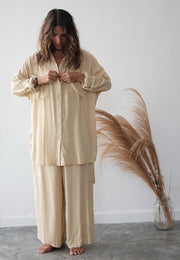 Woman wearing a beige pajama set standing next to a vase with dried pampas grass against a white wall.