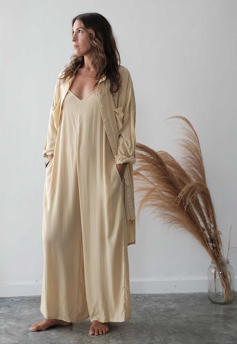 Woman wearing a beige long-sleeve dress standing next to a decorative plant against a white wall.