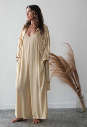 Woman wearing a beige long-sleeve dress standing next to a decorative plant against a white wall.