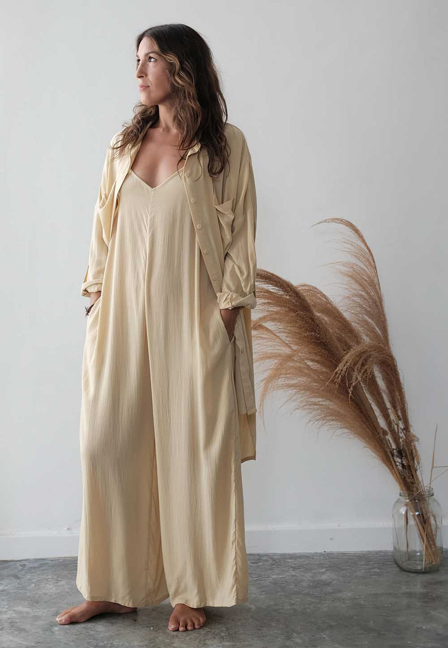 Woman wearing a beige long-sleeve dress standing next to a decorative plant against a white wall.